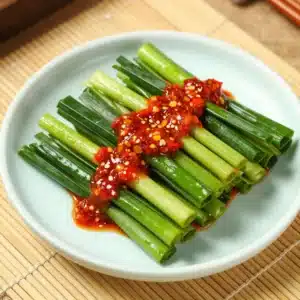 A close-up of Garlic Chive Kimchi (Buchu Kimchi) in a white bowl, showcasing vibrant green chives and red seasoning.