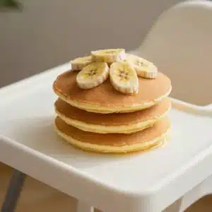 A stack of fluffy banana pancakes topped with sliced bananas and a drizzle of syrup, served on a white plate.