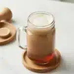 Homemade chai syrup in a glass jar next to a cup of chai latte on a wooden table.