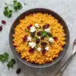 A bowl of creamy pumpkin risotto topped with crumbled goat cheese, dried cranberries, and fresh herbs.