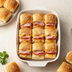 Baked ham and cheese sliders stacked on a tray with melted cheese and glossy golden tops