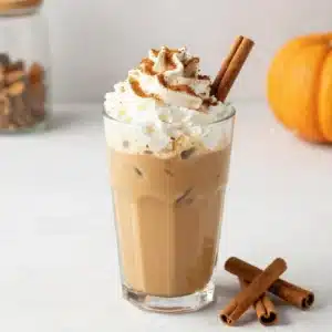A glass of pumpkin spice cold brew topped with creamy foam, cinnamon, and a sprinkle of pumpkin spice on a wooden table.