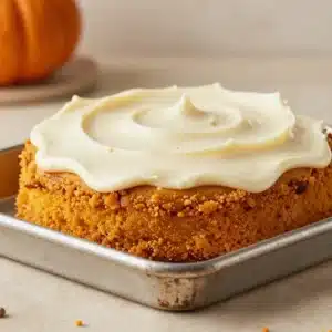 A moist pumpkin sheet cake topped with creamy frosting and a light dusting of cinnamon on a white baking tray